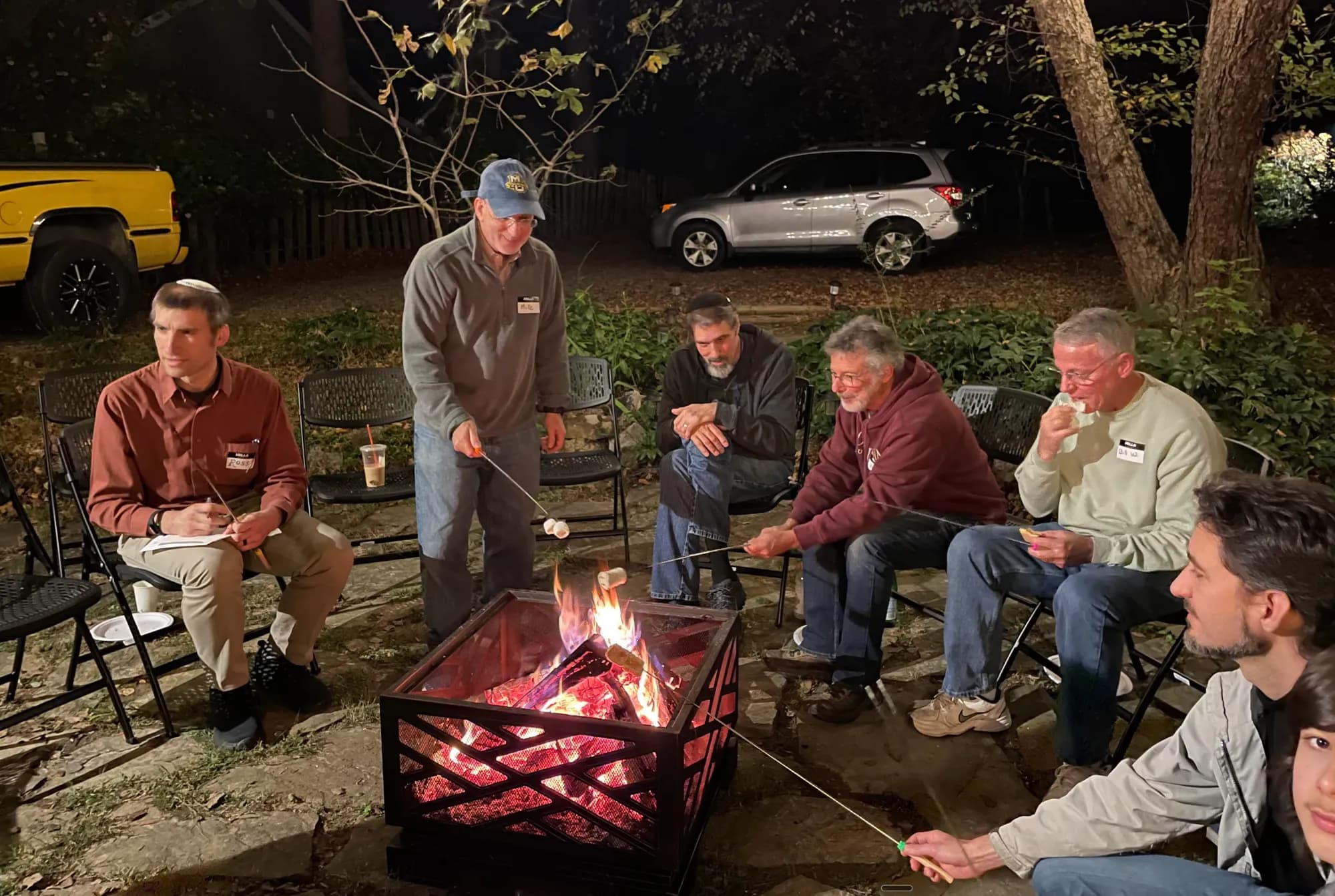 Men of the congregation gathered around a fire pit at night, roasting marshmallows