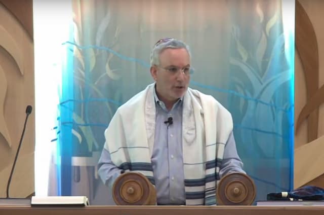 Reb Mike Kaufman, wearing a tallit, teaching from the bimah with the Torah scroll open before him